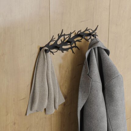 Μία μεταλλική κρεμάστρα μαύρη σε σχήμα κλαδιού τοποθετημένη σε ξύλινο τοίχο με κρεμασμένα ένα γκρι παλτό και ένα κασκόλ